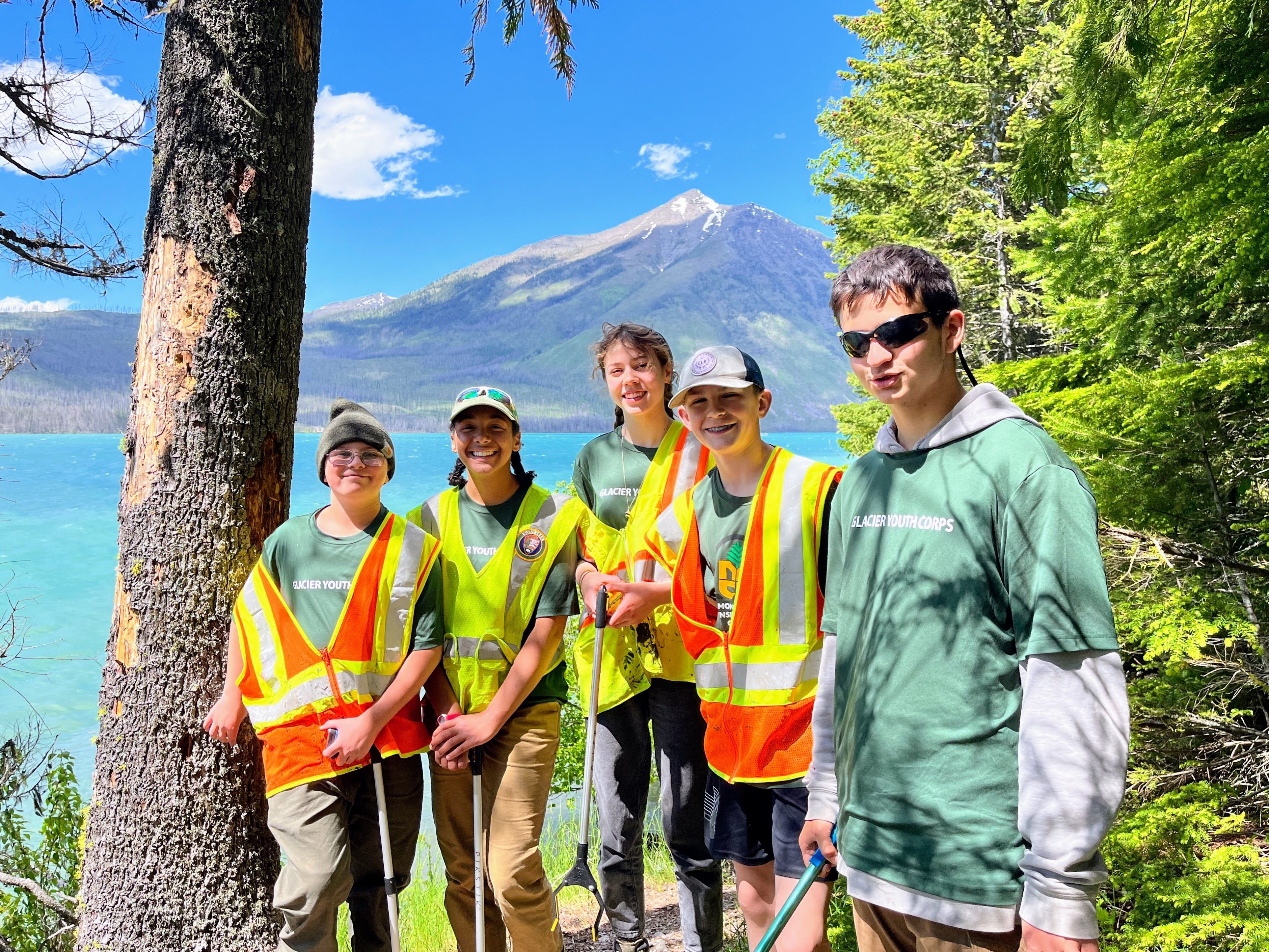 A group of teens wearing safety vests pose in front of a bright blue lake.