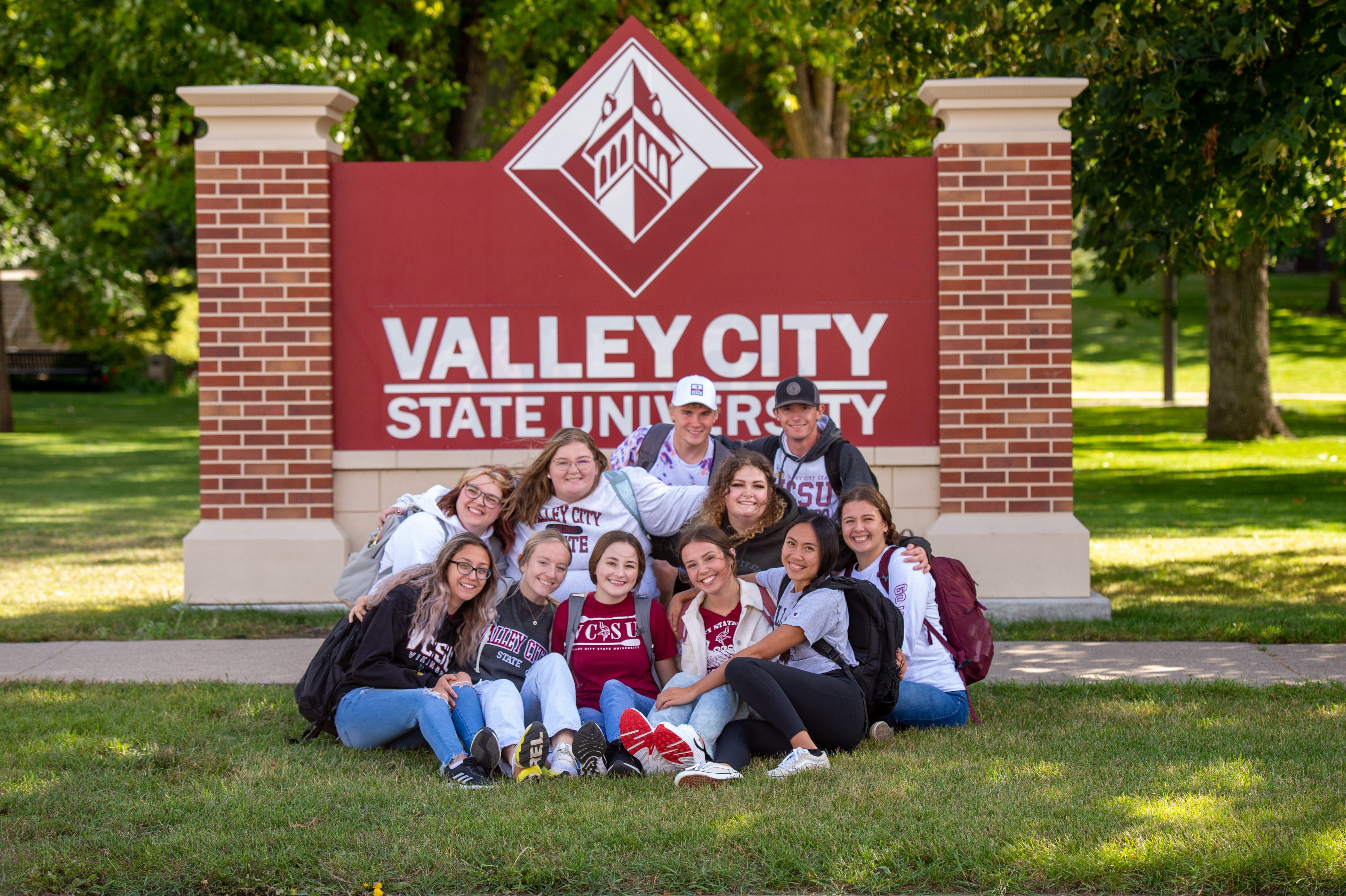 Group of students huddle up on the ground in front of the VCSU sign smiling at the camera