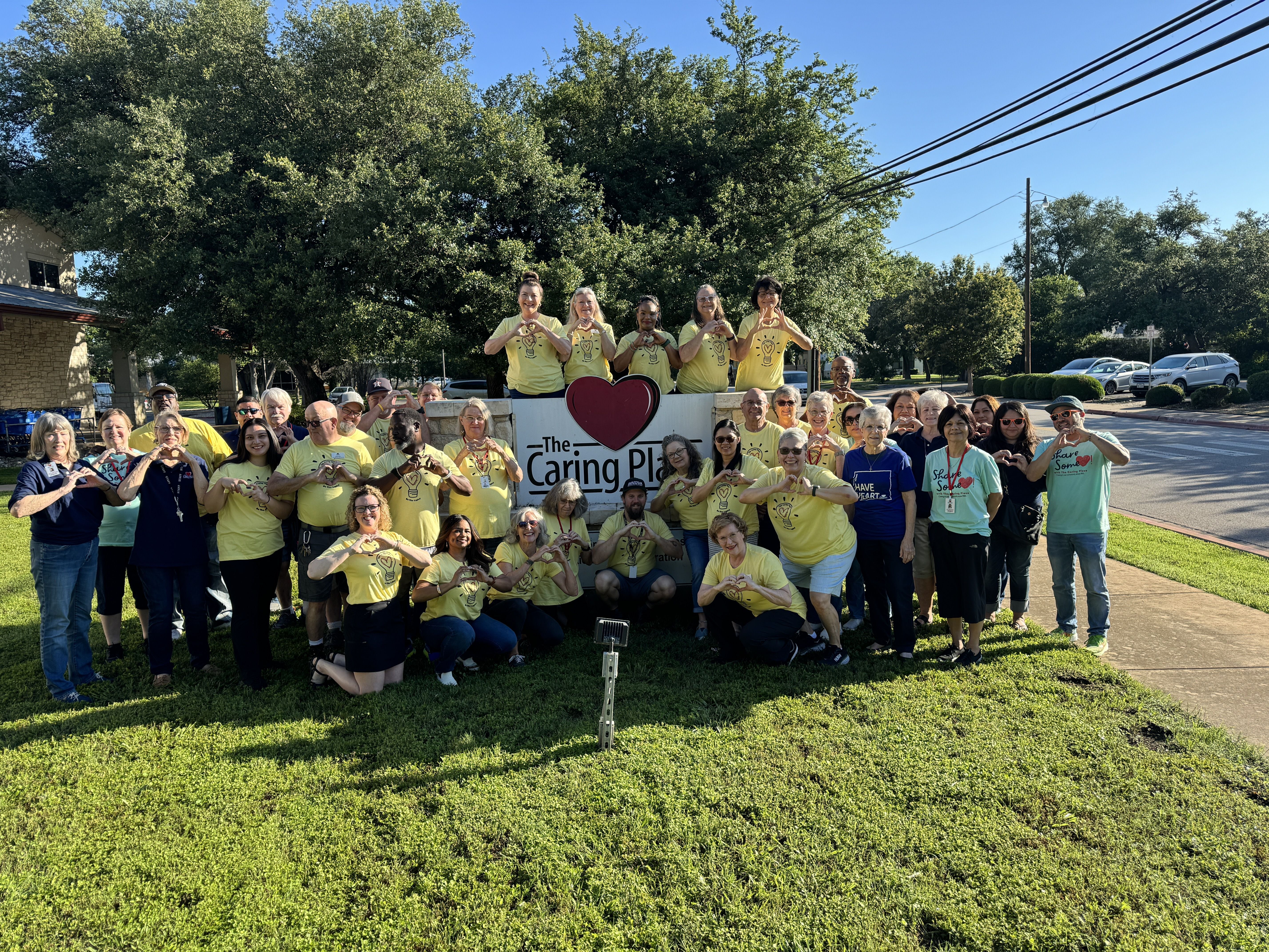 A group of volunteers and staff from The Caring Place gather at the Headquarters Sign making heart shaped gestures with their hands celebrating and honoring the caring love of The Caring Place