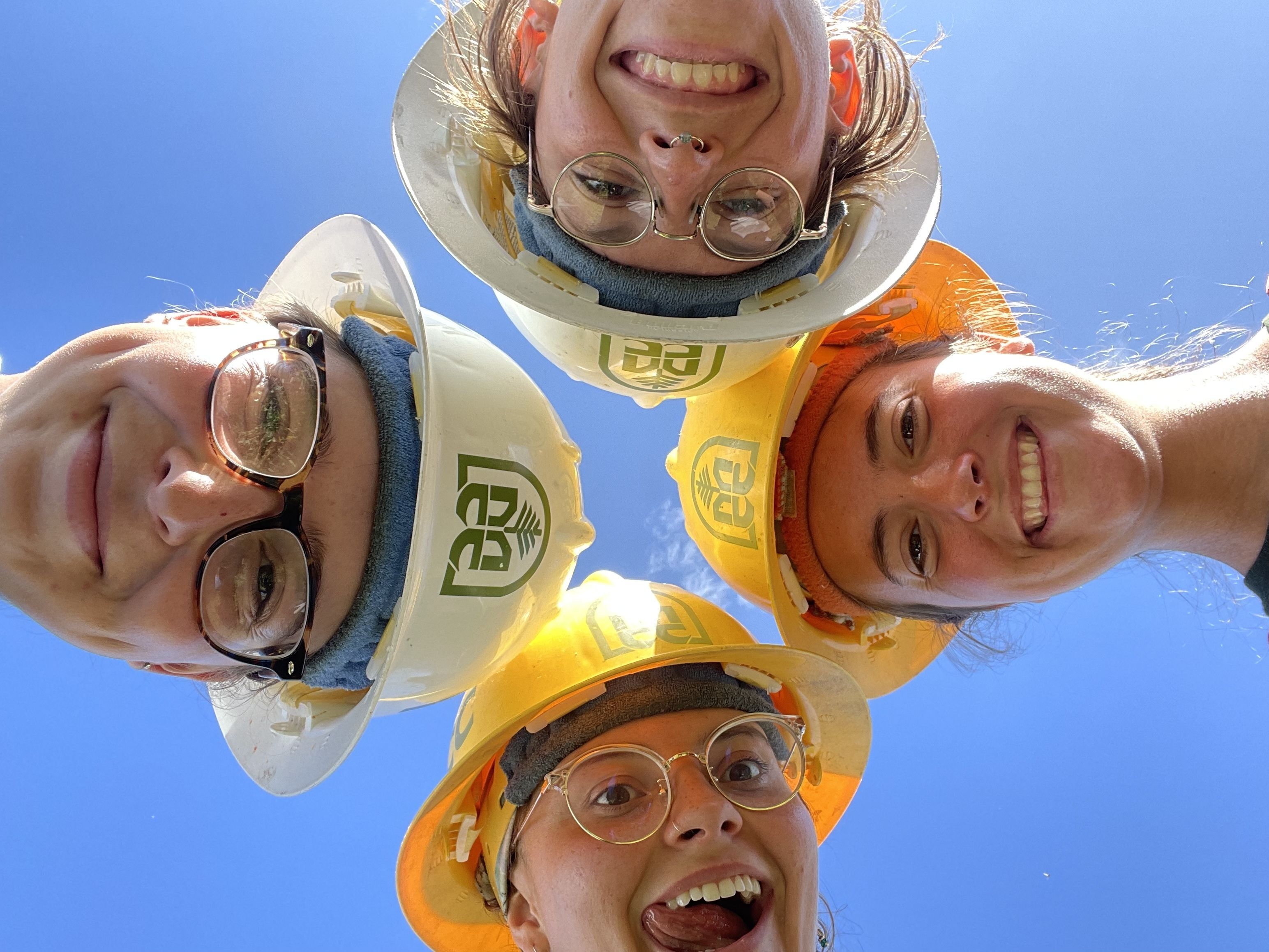 Four crew members wearing hard hats cluster in a circle above the camera, and against a blue sky. They are smiling and laughing.
