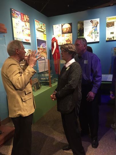 Man in a nehru jacket talking to a couple standing in front of a display of 1960 sporting equipment in the Smoky Hill Museum's 1960 exhibit.