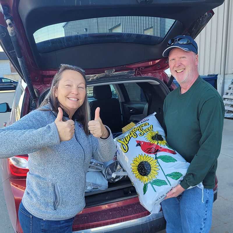 Blair Laddusaw holding a bag of sunflower seeds for Amy Campagna, who is smiling and giving a thumbs up sign in front of her car's trunk