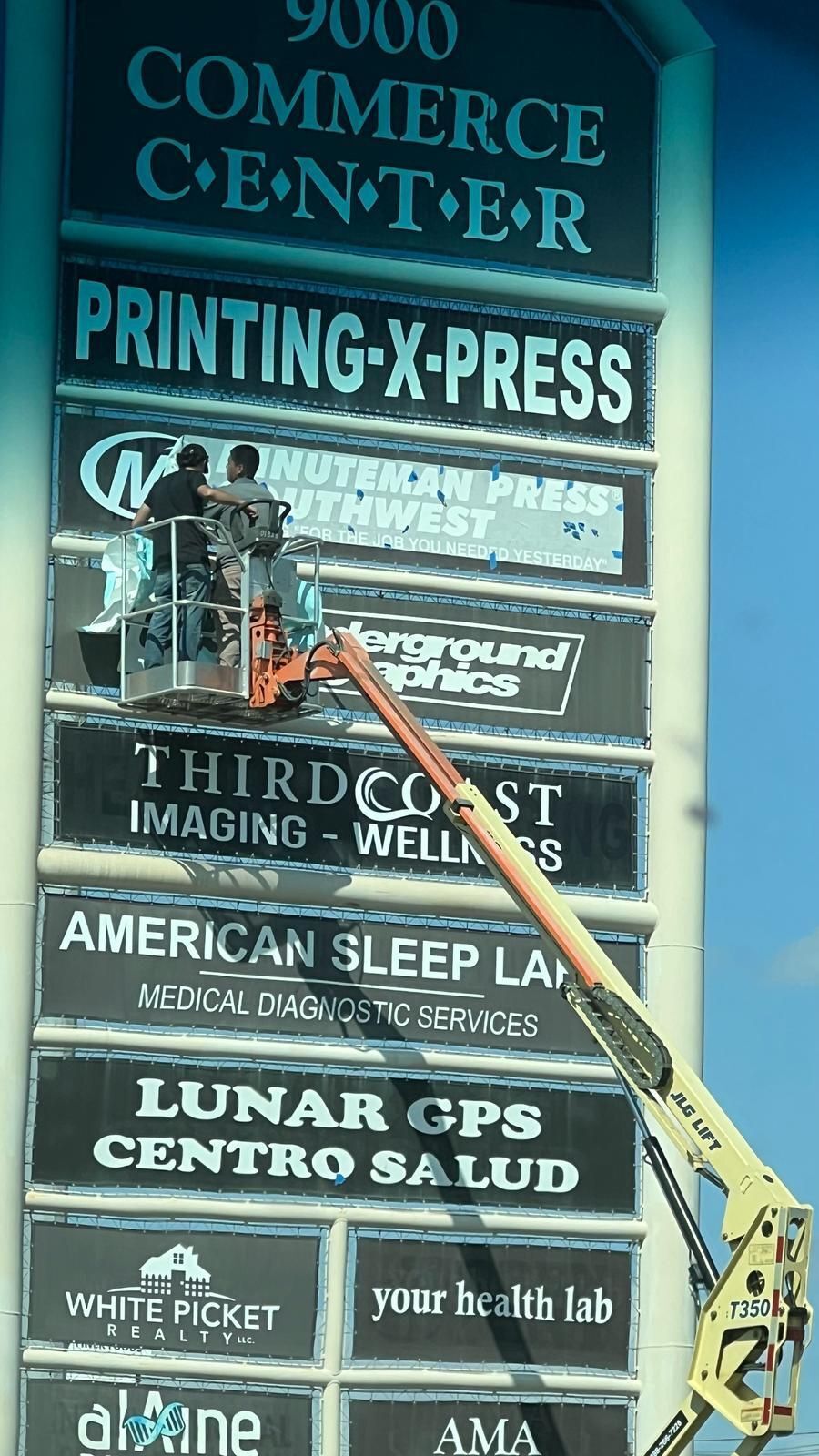 Two technicians from the install team using a boom lift to mount business signage panels on a multi-tenant pylon sign at 9000 Commerce Center under daylight conditions.