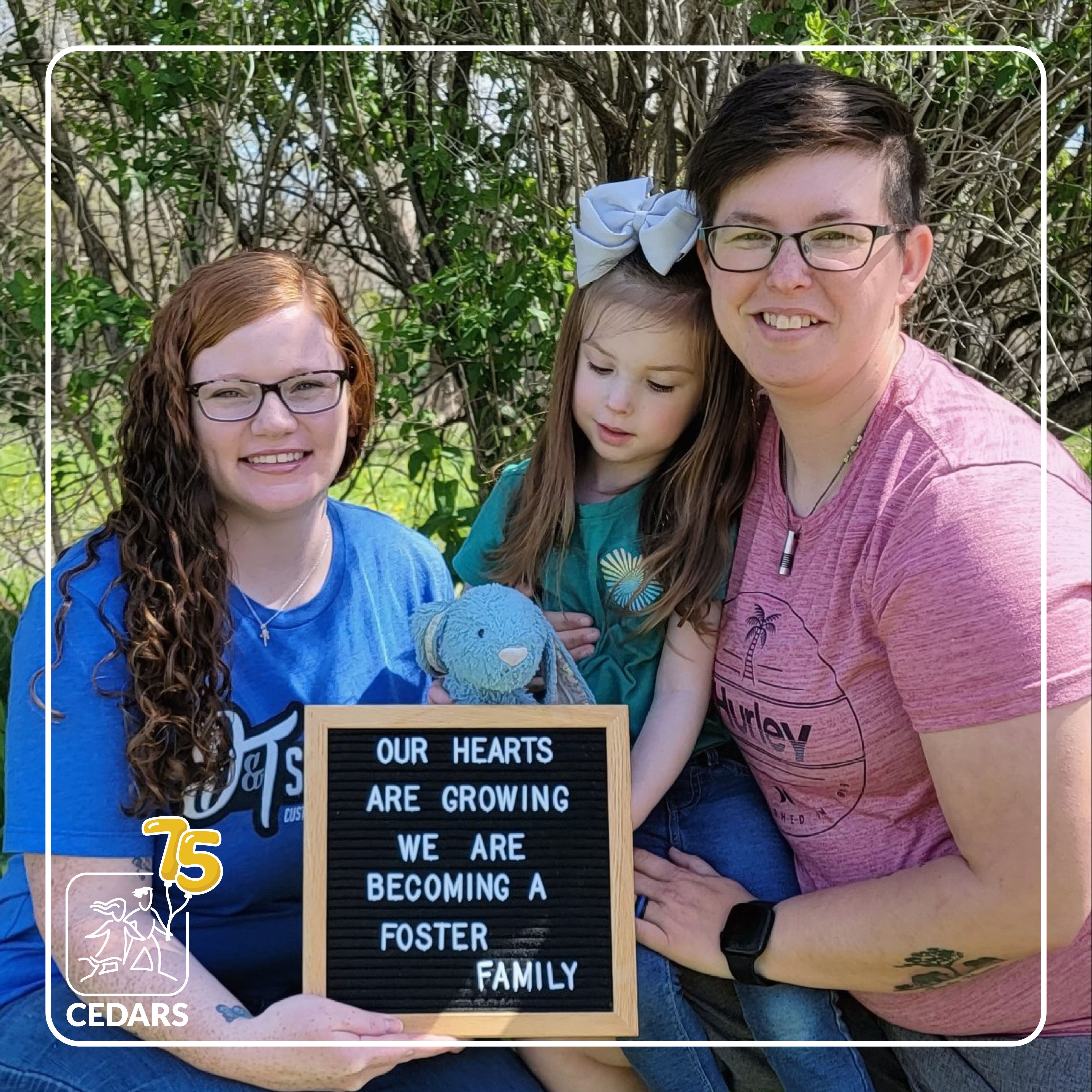 Photo of a family together outdoors. The two moms kneel down holding their young daughter between them and displaying a sign that reads: "Our hearts are growing. We are becoming a foster family."