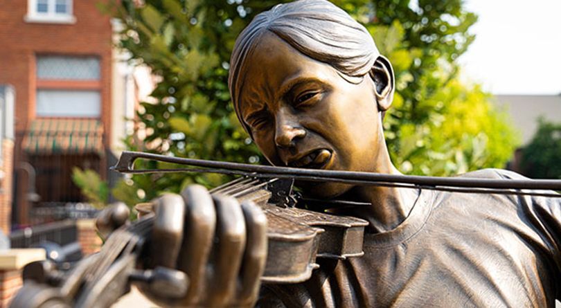 A bronze sculpture of a Tom girl practicing on her violin with her hound puppy laying by her.  #15 Gotta Practice First by Sherri Treeby & Lee Leuning
