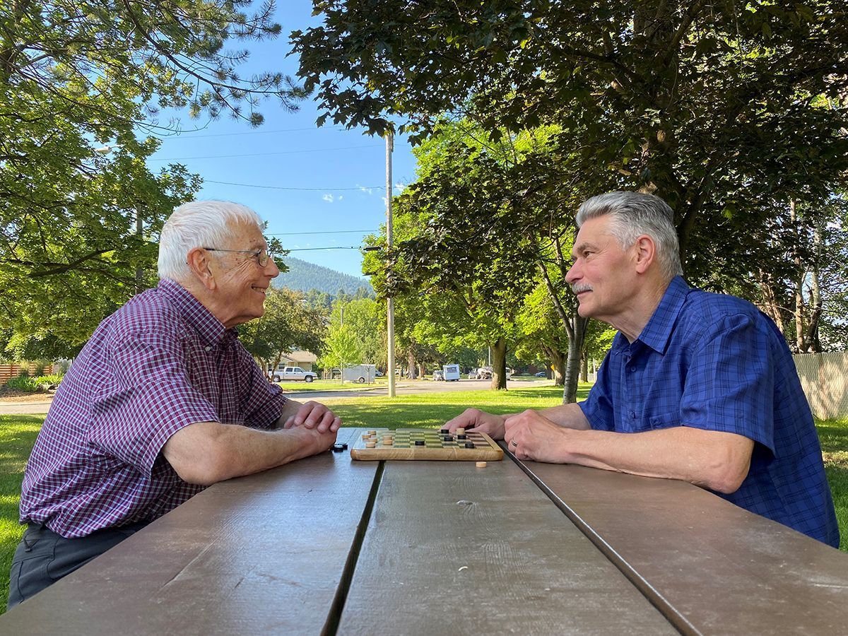 Two male older adults sitting at a park picnic table on a summer day playing checkers