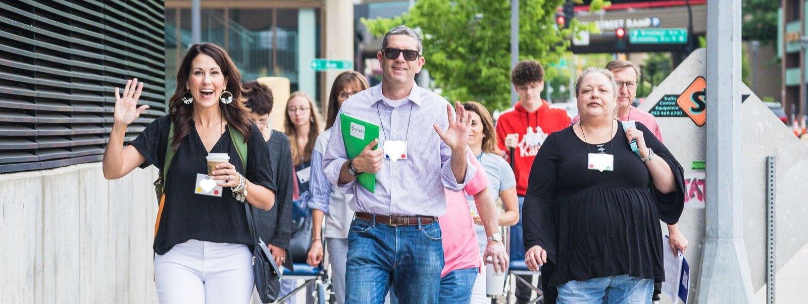 People are walking down the street toward the camera. The three in the front, a woman, man, and woman. are all smiling. The woman on the left is waving.