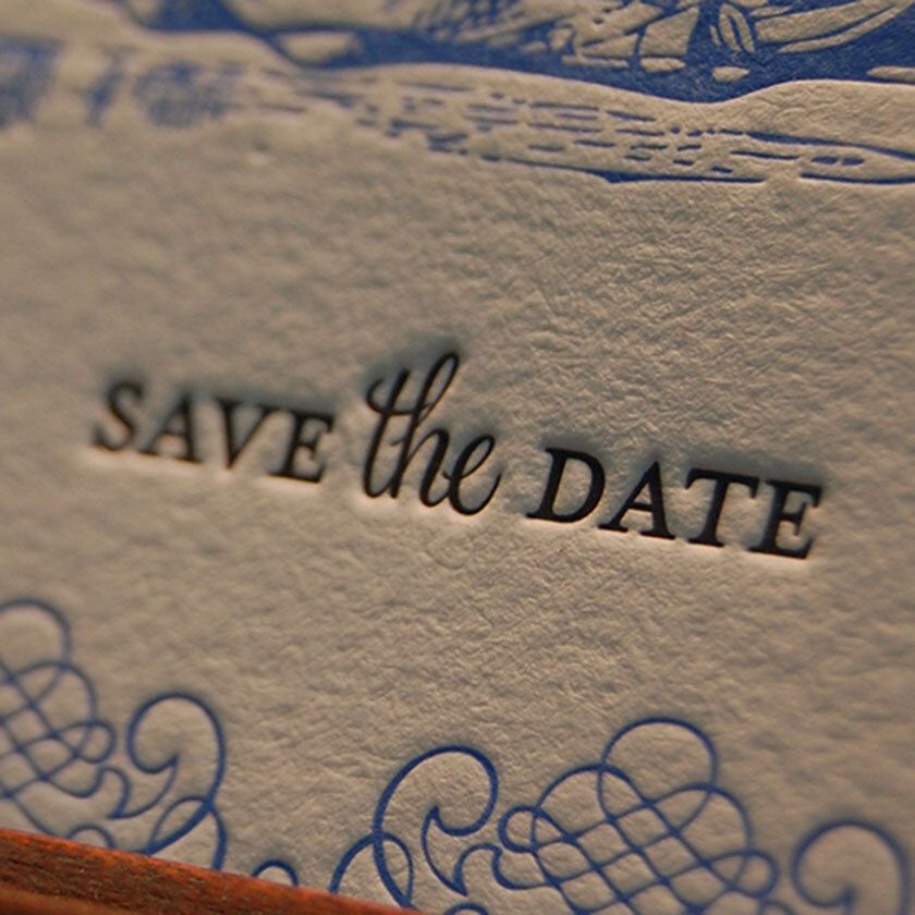 Close-up of embossed text "SAVE the DATE" on textured paper with blue decorative swirls.