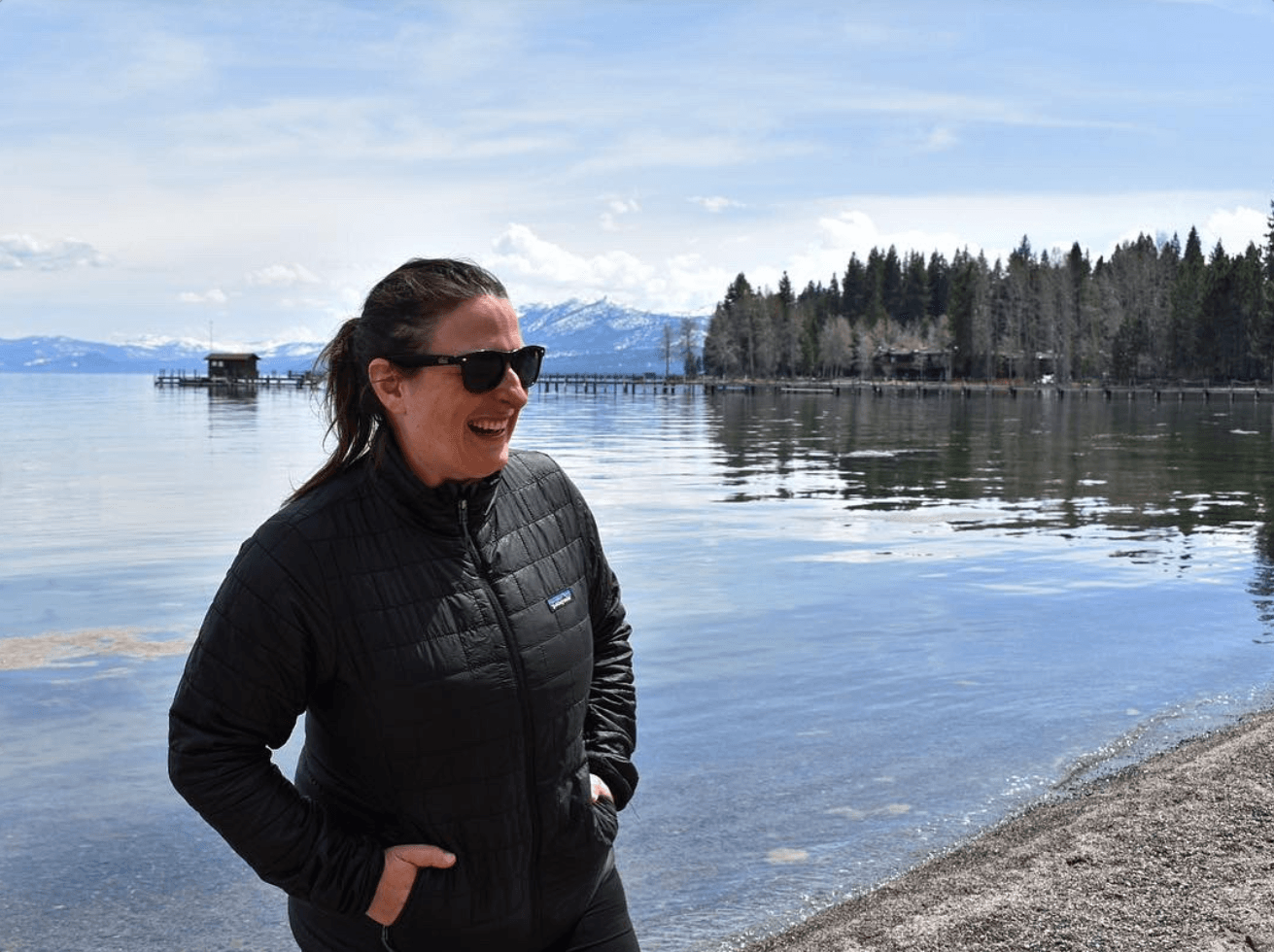 A smiling woman stands in front of a lake, hands in the pockets of her black jacket.