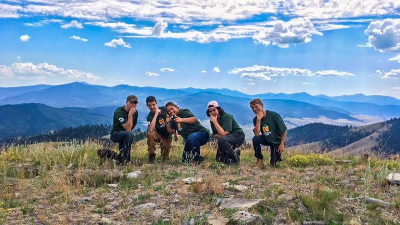 [Image Description: Five MCC Crew members kneel in a pose on a mountaintop field.]