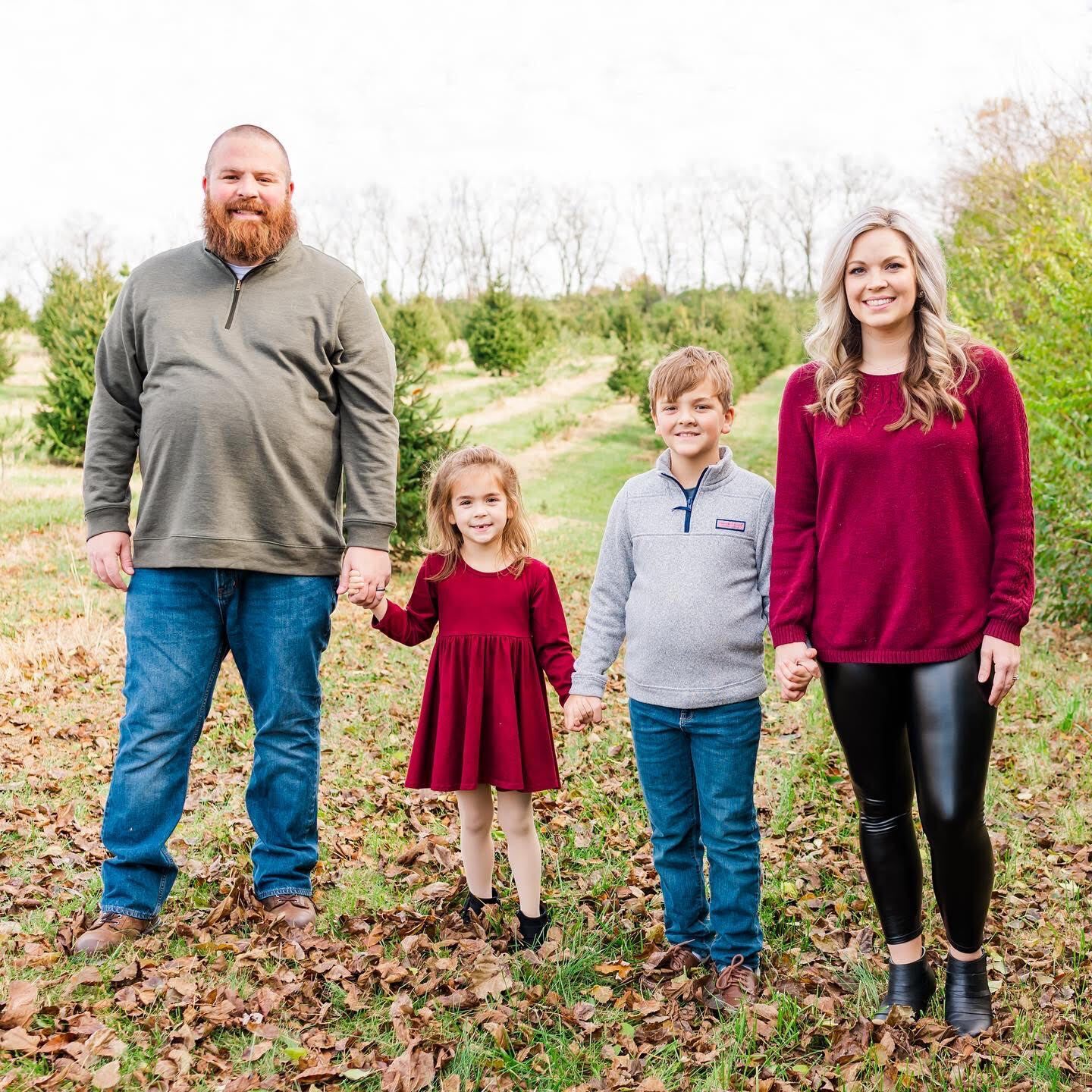 Family photo taken in an open field with evergreens in the distance. L-R: Dad with mustache and beard, jeans and long sleeved shirt holds hand of young girl next to him. She has blond hair and is wearing a red dress, holding the hand of her older brother 