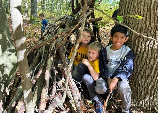 3 kids, the one in front grinning widely, try to hide in a small wood fort made of sticks leaning against a large tree