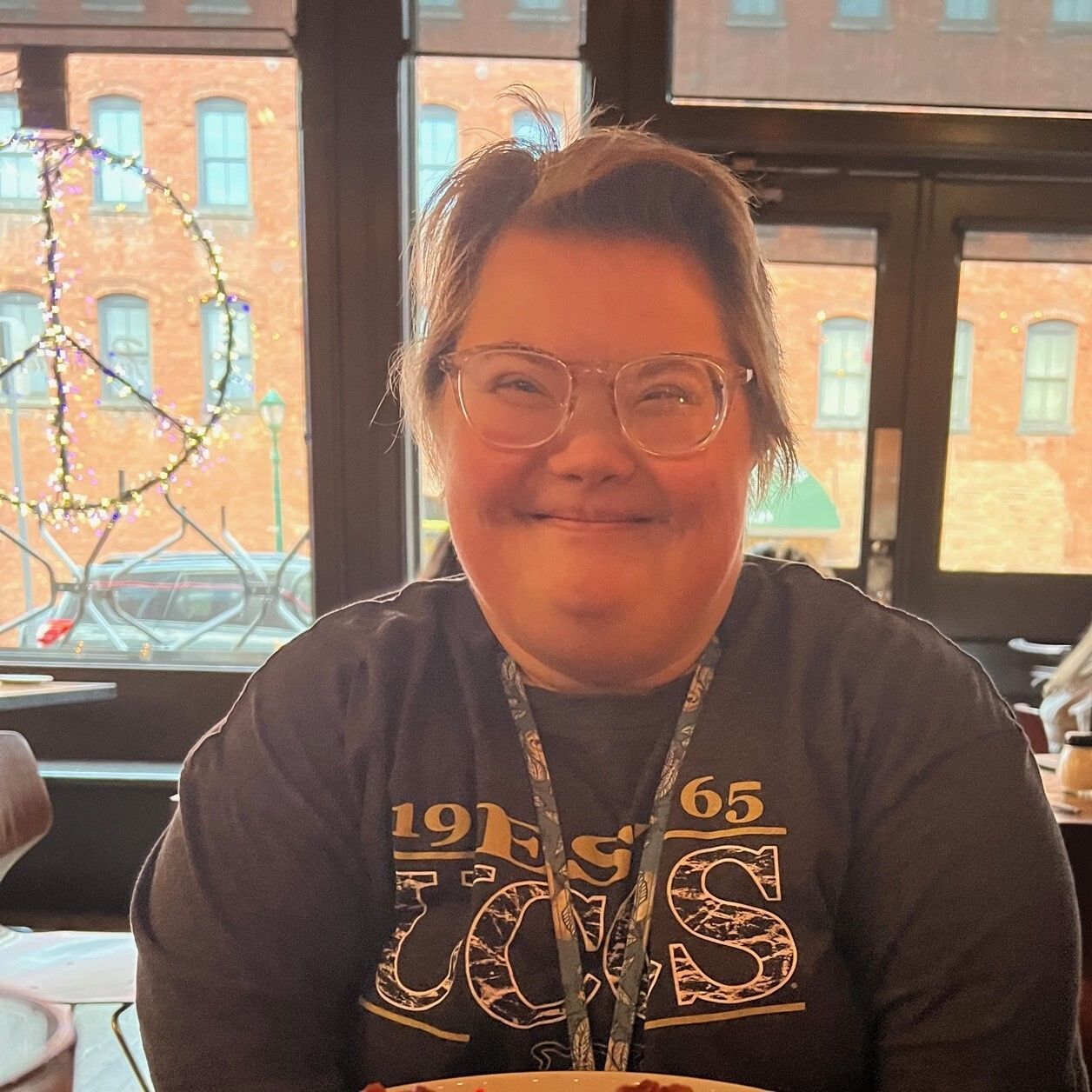 Georgie smiling with glasses on and a UCCS shirt. A peace sign hangs in the background. 