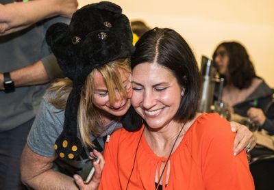 One woman in an orange shirt is sitting and smiling. Another woman wearing a bear hat is standing behind her hugging her shoulders.