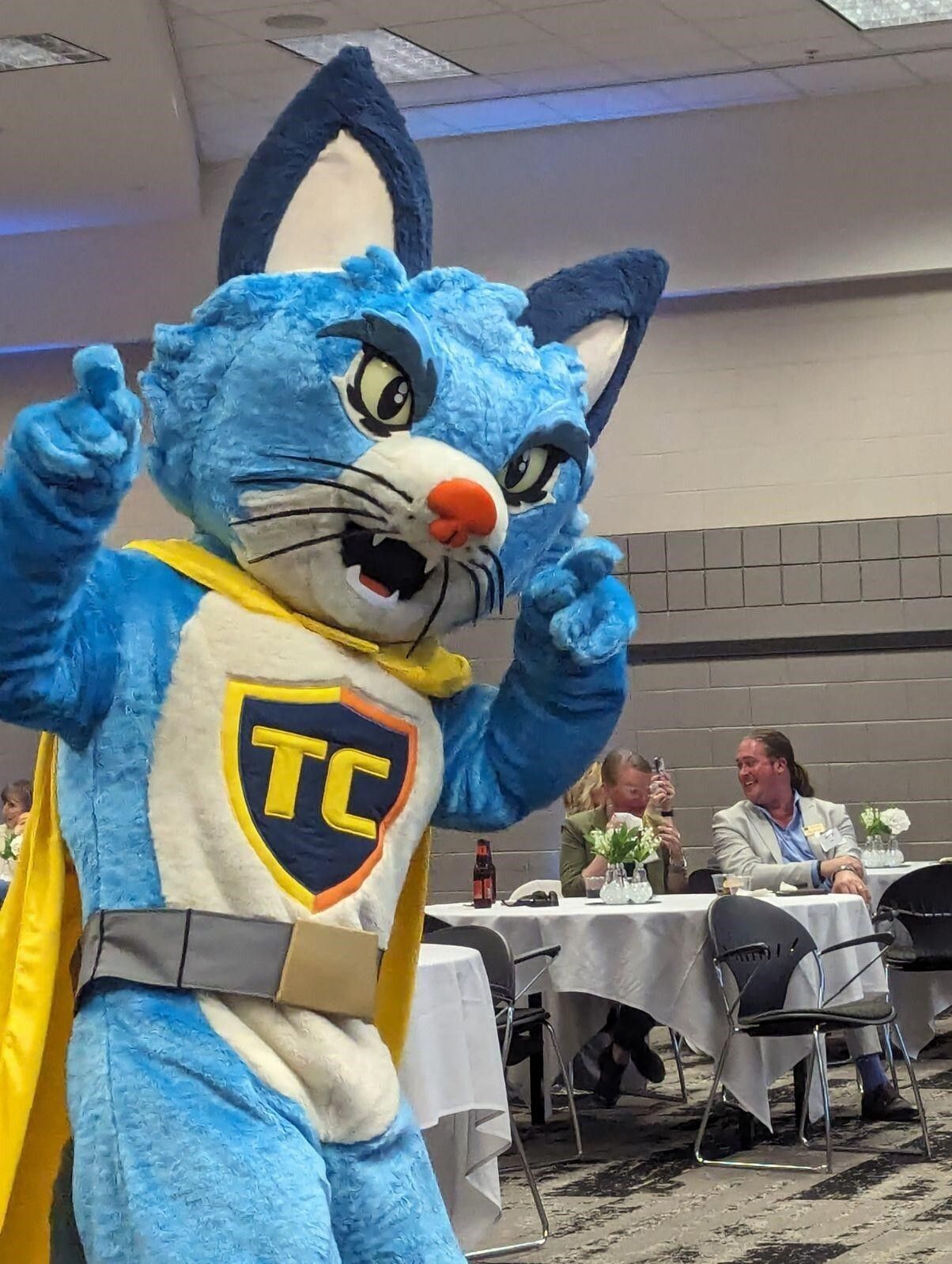 A person in a blue cat costume that has a "TC" crest on its chest and is wearing a yellow cape