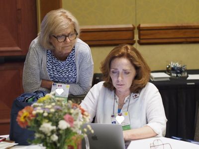 A woman sits at a laptop. Another woman stands behind her, leaning forward.