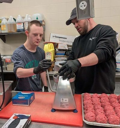 2 men sit at a table where 1 helps the other to squeeze green paint out of a bottle