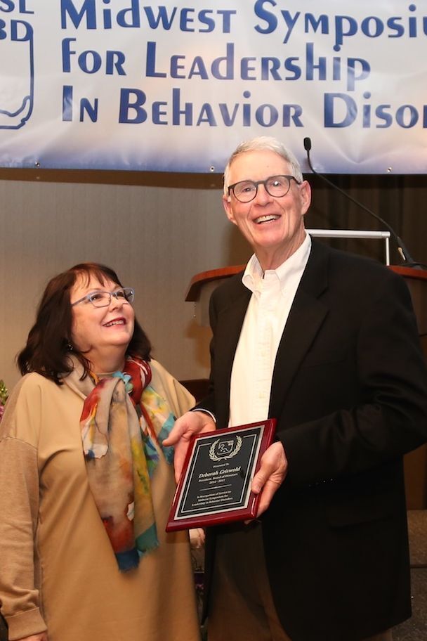 At the Midwest Symposium for Leadership in Behavior Disorders, a tall older man in a black blazer and white shirt holds a red-framed award plaque while a smiling woman in a tan top and colorful scarf looks up at him beneath the event banner.