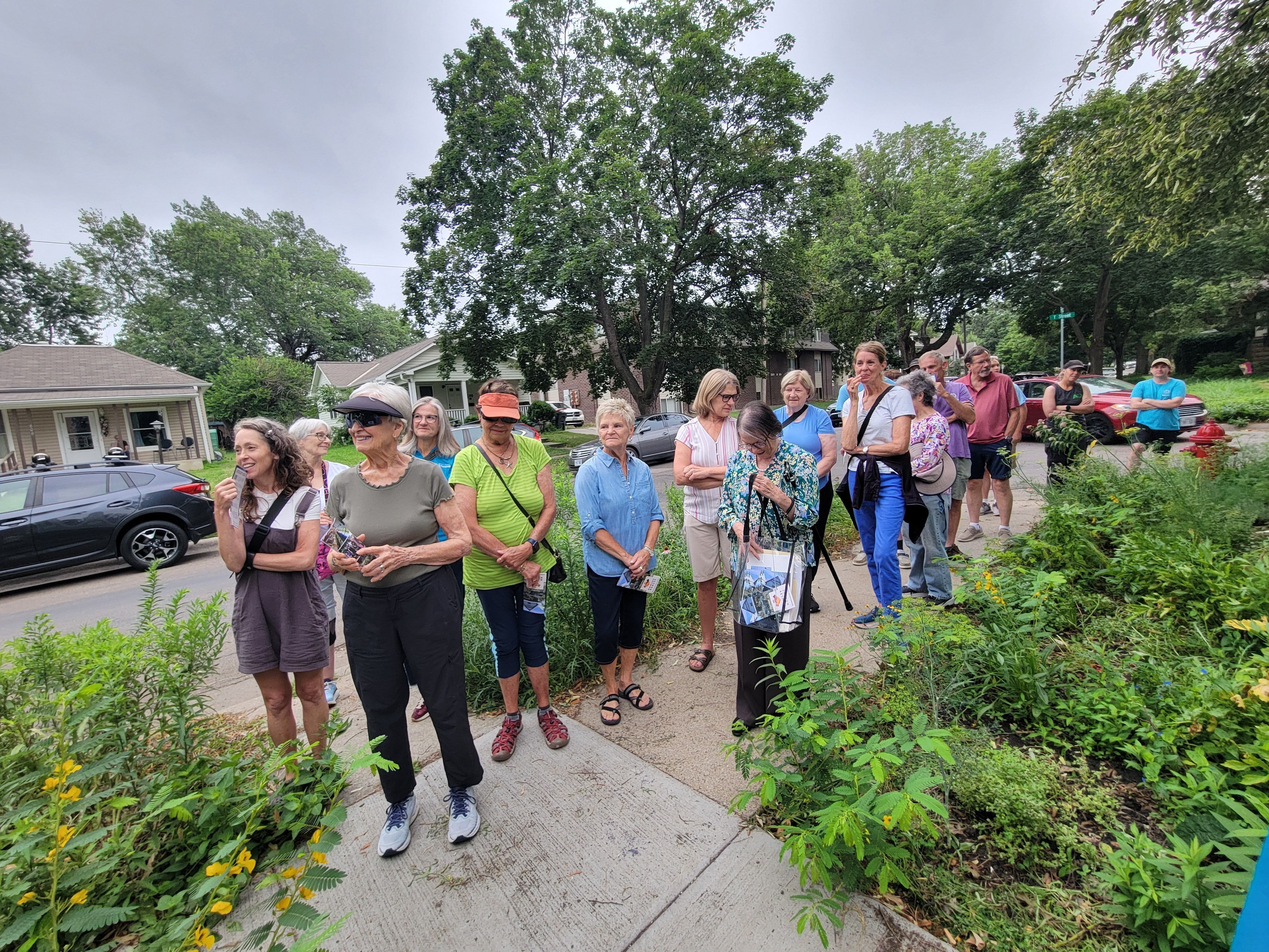A group of people gathered on a residential sidewalk surrounded by native plantings during an outdoor garden tour on an overcast summer day.