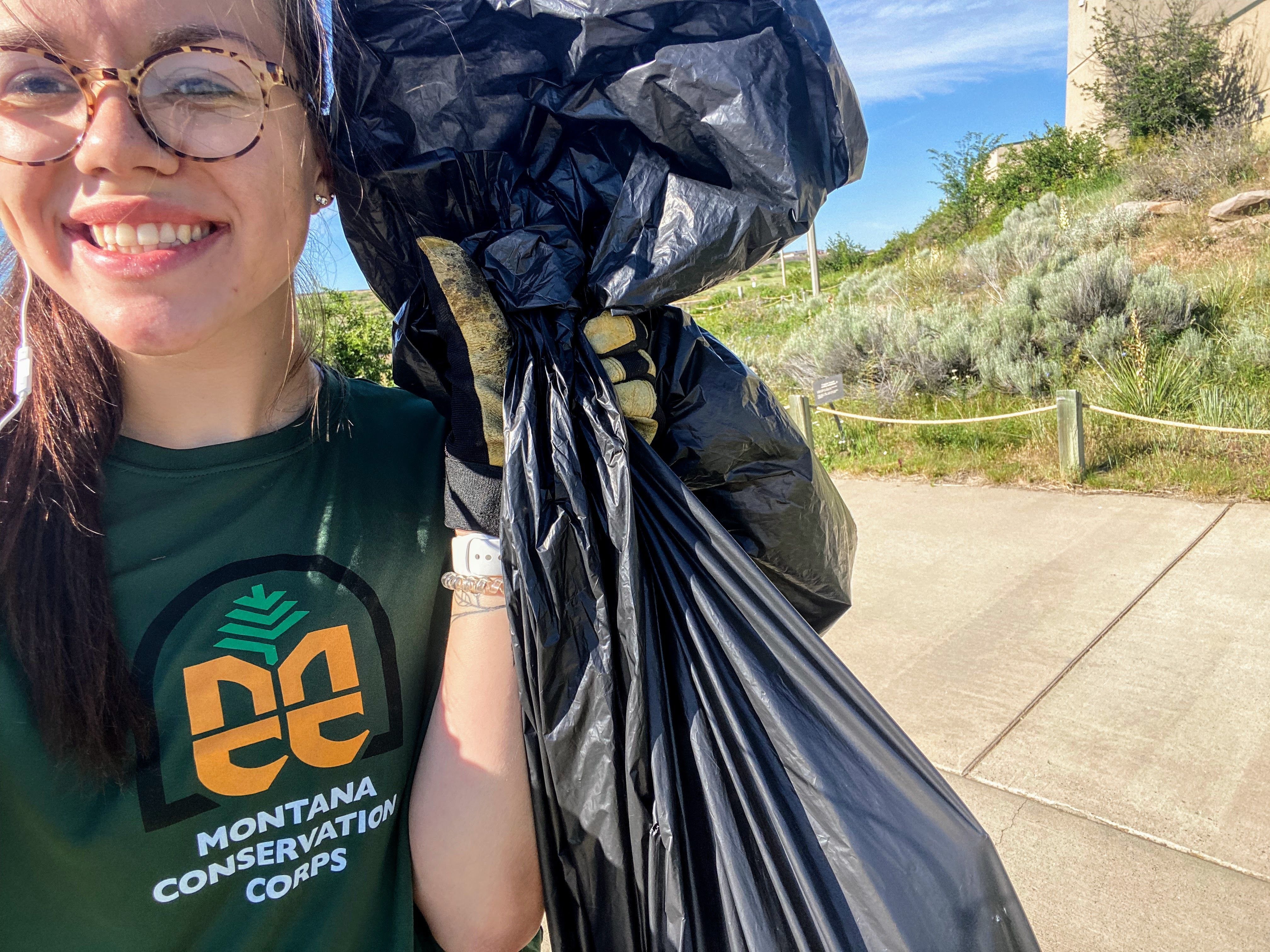Lily holds a trashbag up next to her face and smiles at the camera. She is wearing a green shirt with an MCC logo.