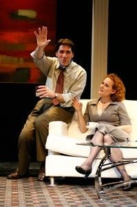 Nicholas is wearing business casual attire and raising one of his hands while he's talking. He's sitting on the armrest of a couch and Marilee is sitting on the couch and listening to Nicholas. 