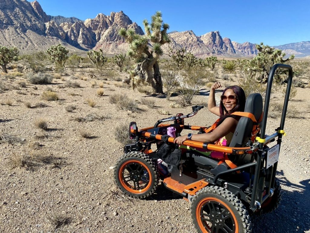 A woman flexes her strong arm muscles in excitement as she rides a TerrainHopper all terrain wheelchair down a desert trail at Red Rock Canyon National Conservation Area. 