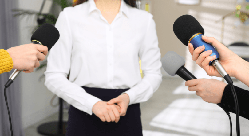 Image shows young woman with brown braided hair using a video camera to record an interview of a woman while surrounded by other people