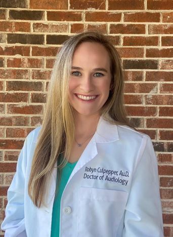 Photo of Robyn,  a white woman with blue eyes, and blonde hair smiling. She is wearing a white Doctor of Audiology coat.