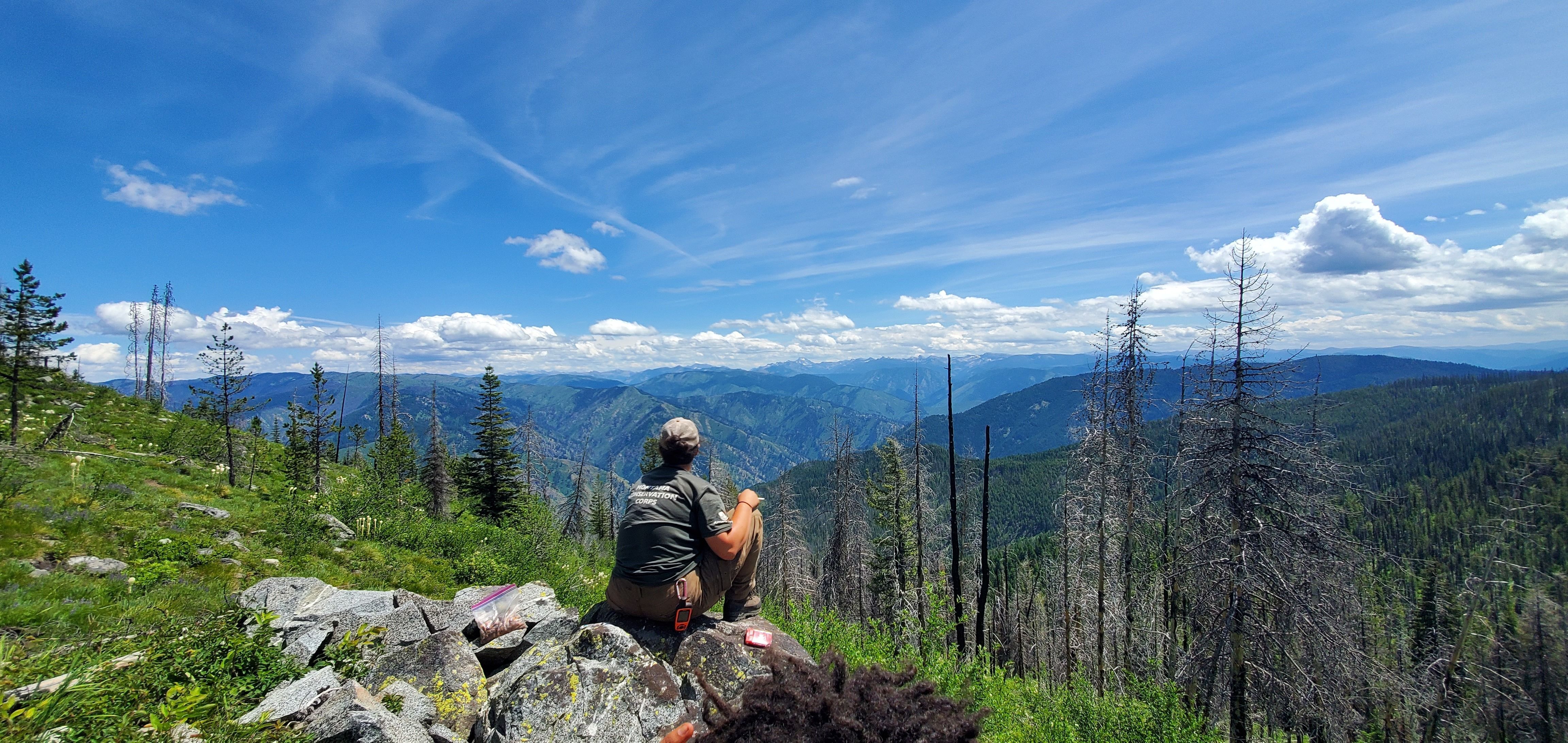 A person wearing an MCC shirt sits on a rock, overlooking a valley below with mountains and blue sky in the distance.