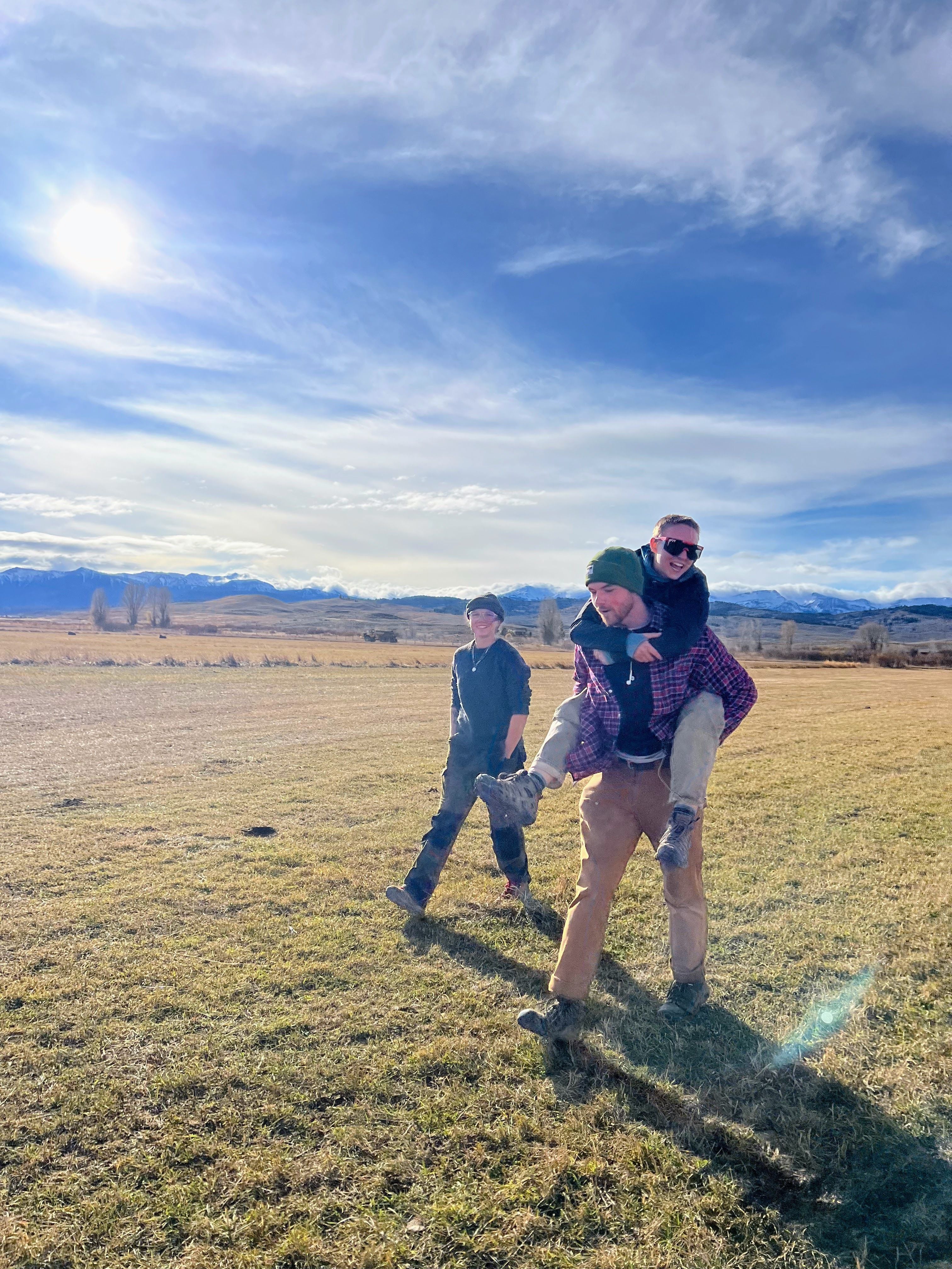 A crew member giving a piggy back to another member across a field, with another field crew member walking in the background.