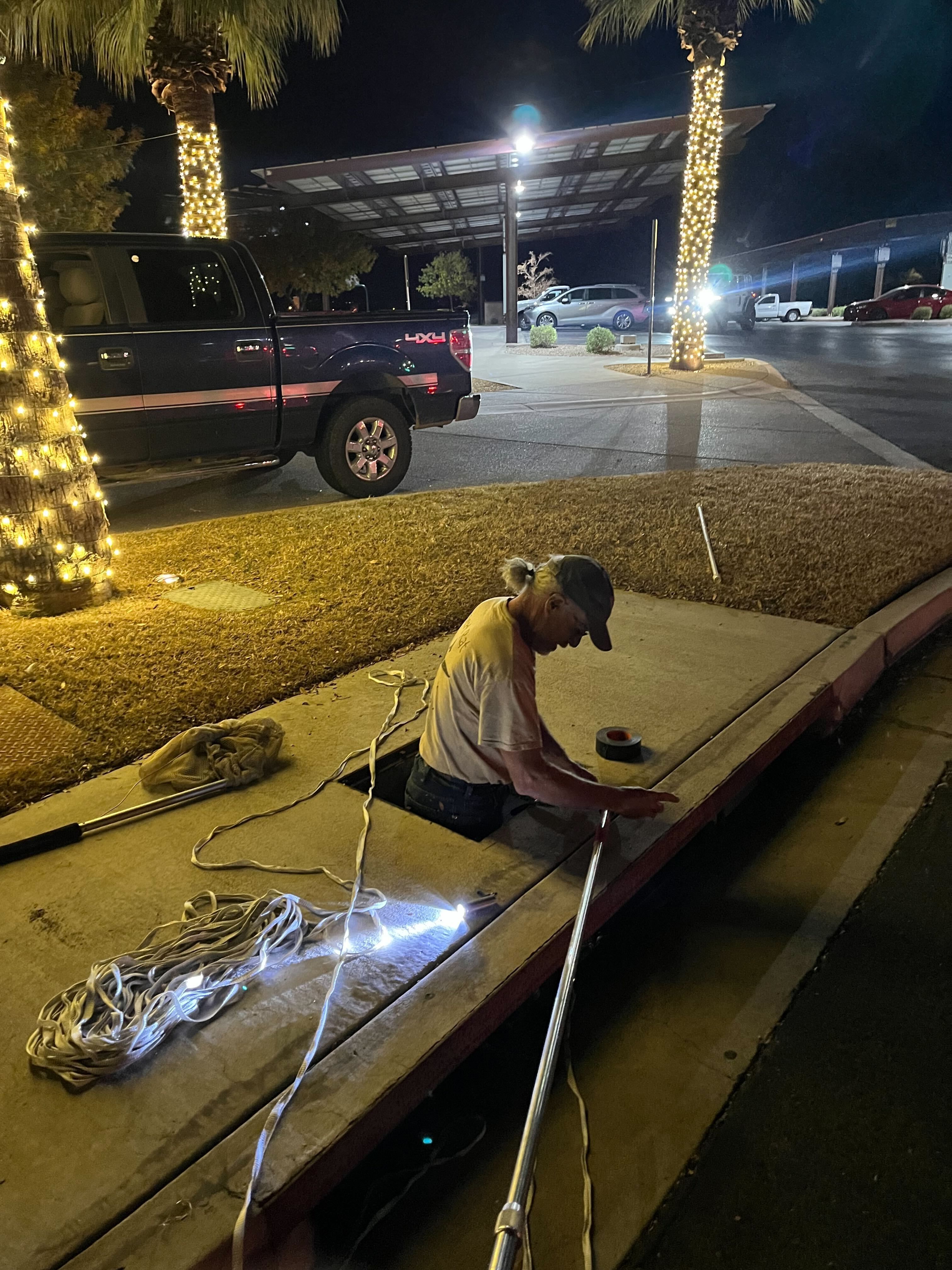 Rescuer Paul is half-in, half-out of a vertical storm drain entrance at night. He's lit by twinkling holiday decorations in the background. He is deep in thought, brainstorming how to save the raccoon's life.