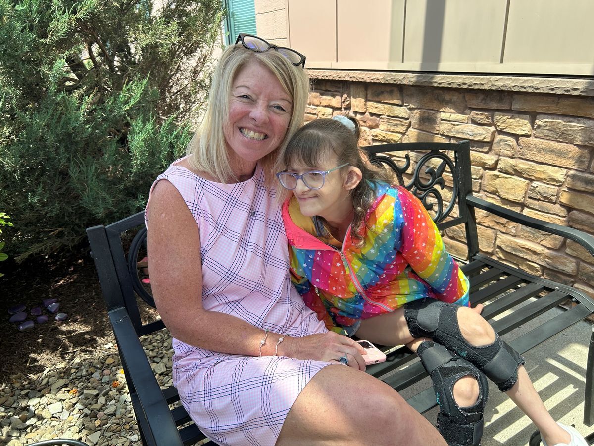 A woman sits on a bench smiling and facing a camera and a young girl leans into her.