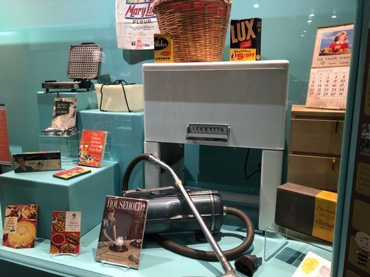 Exhibit case at the Smoky Hill Museum showing lots of 1950s items used in the home including an Electrulux canister vacuum cleaner, Tupperware container and waffle maker.