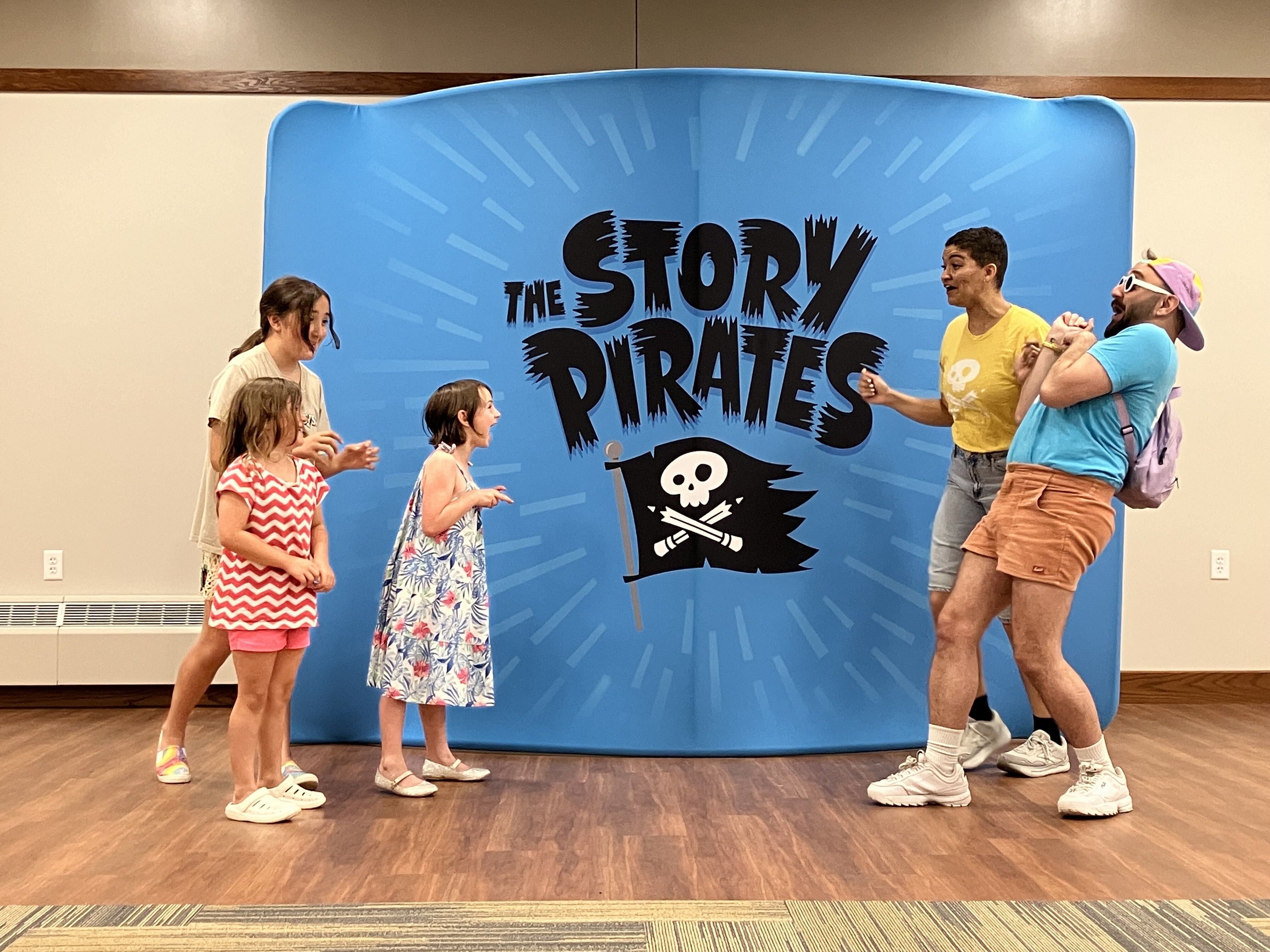 Three children stand excitedly in front of a bright blue "Story Pirates" backdrop, interacting with two animated adult performers in colorful outfits. The performers are playfully reacting to the kids, creating a fun and engaging moment from the Story Pir