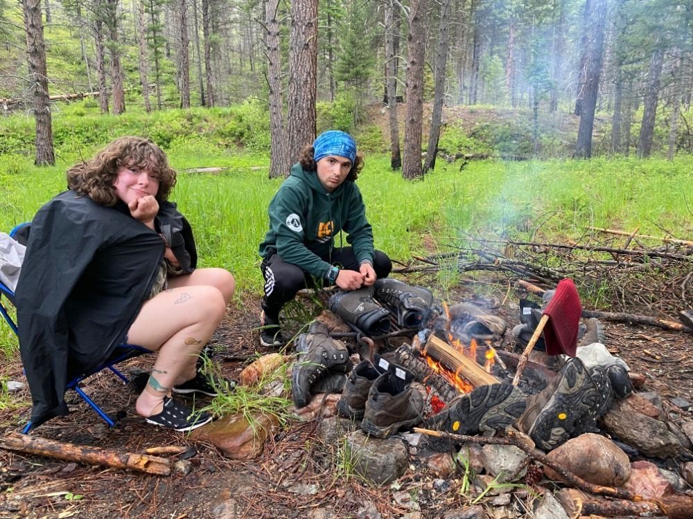 Two people sit huddled around a fire. Many pairs of boots drying out ring the fire.