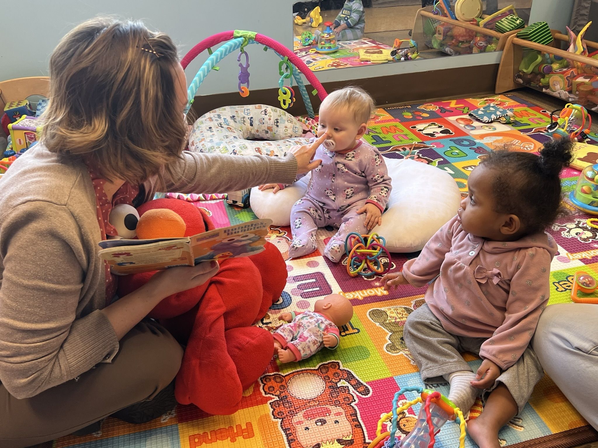 A lady sits on the floor of a classroom with two infants. There are toys surrounding the kids as the lady reads them a book while tapping the nose of one of the infants.