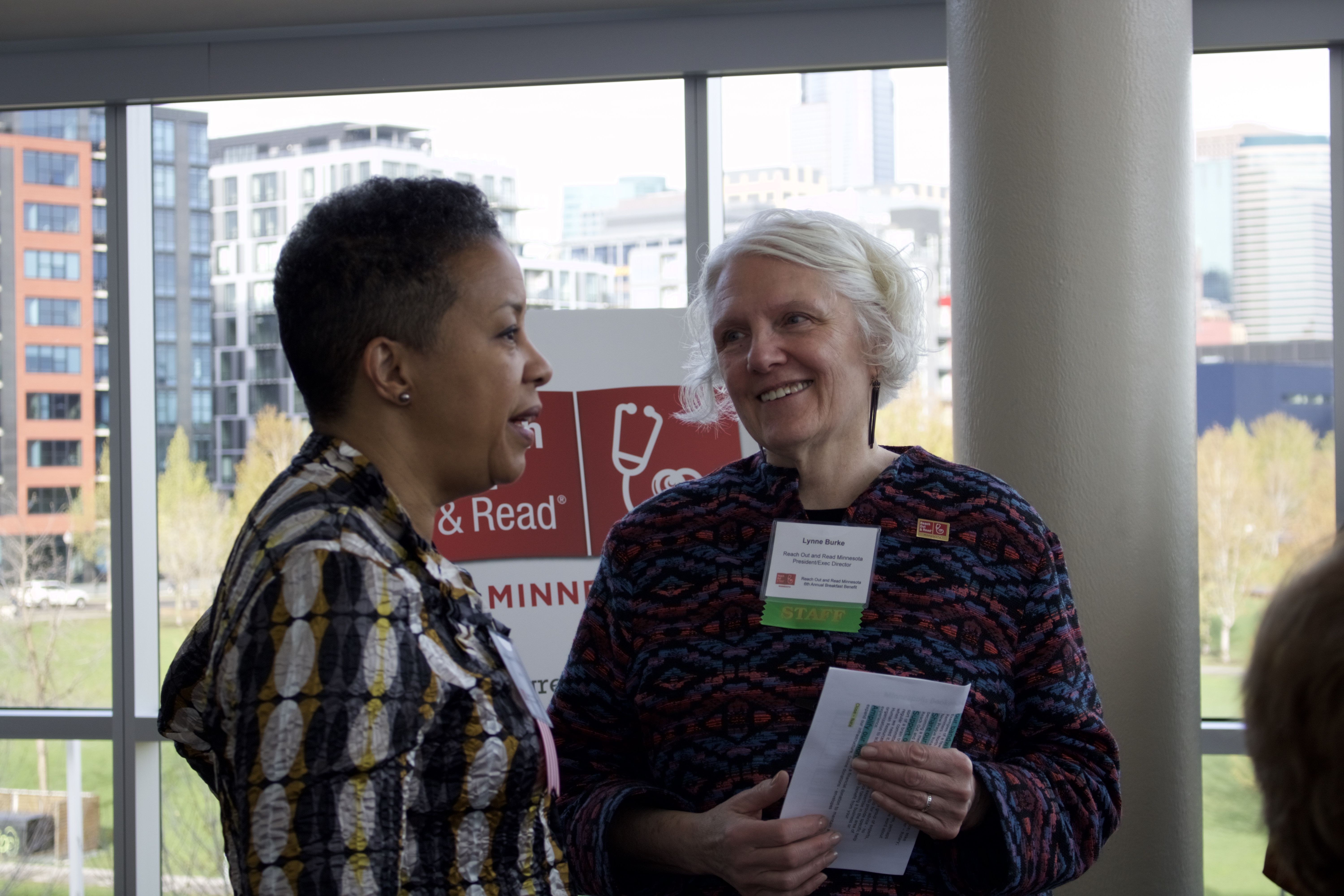 Two women talk in front of a Reach Out and Read Minnesota printed sign