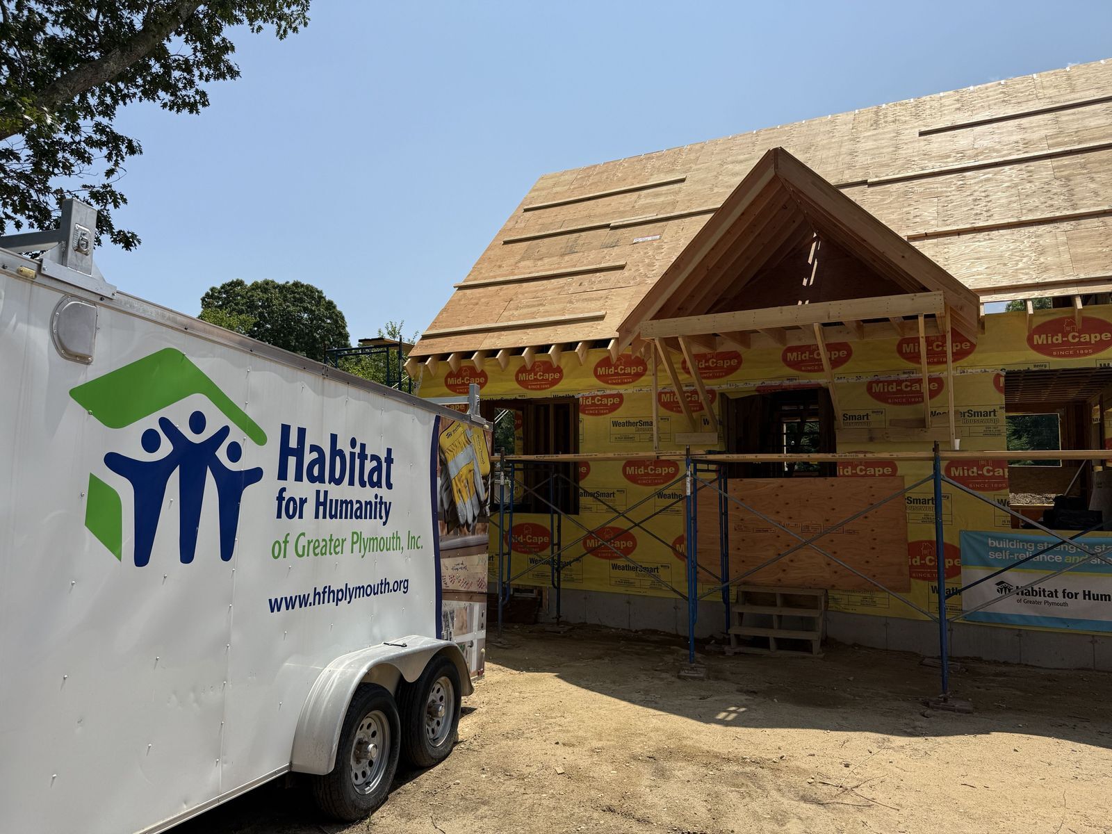 Photo of home build that is fully framed with exterior walls and roof, next to white tool trailer with "Habitat for Humanity of Greater Plymouth" logo