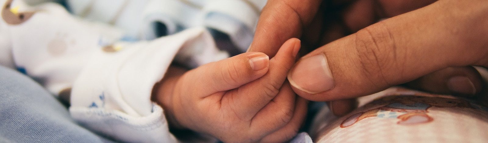 Closeup photo of a mother holding her baby's tiny hand between her index finger and thumb. The baby's arm is in a white sleeve with small animals on it and enters the photo from the left. The mother's hand enters the photo from the right.