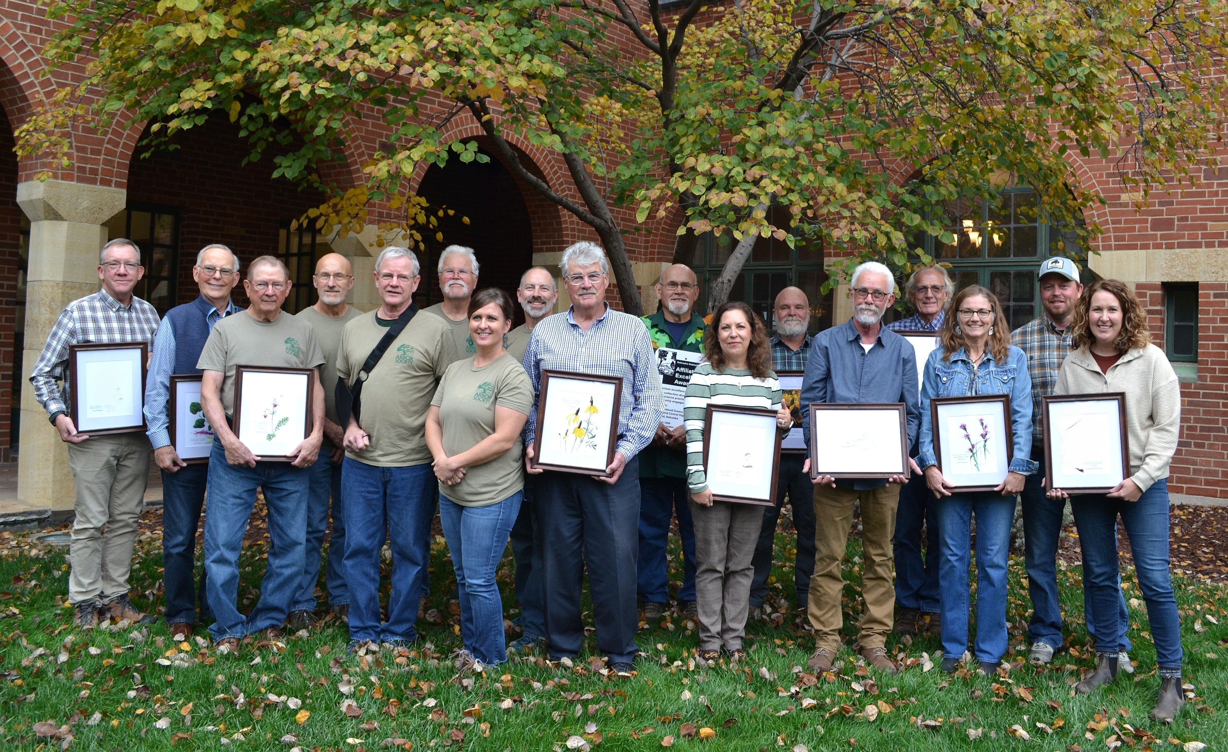 A large group of NSA Award winners holding their framed awards in front of a tree in a courtyard.