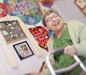 Elderly woman PSRS supports wtih her walker, smiling in front of a wall in her bedroom covered with photos and decorations