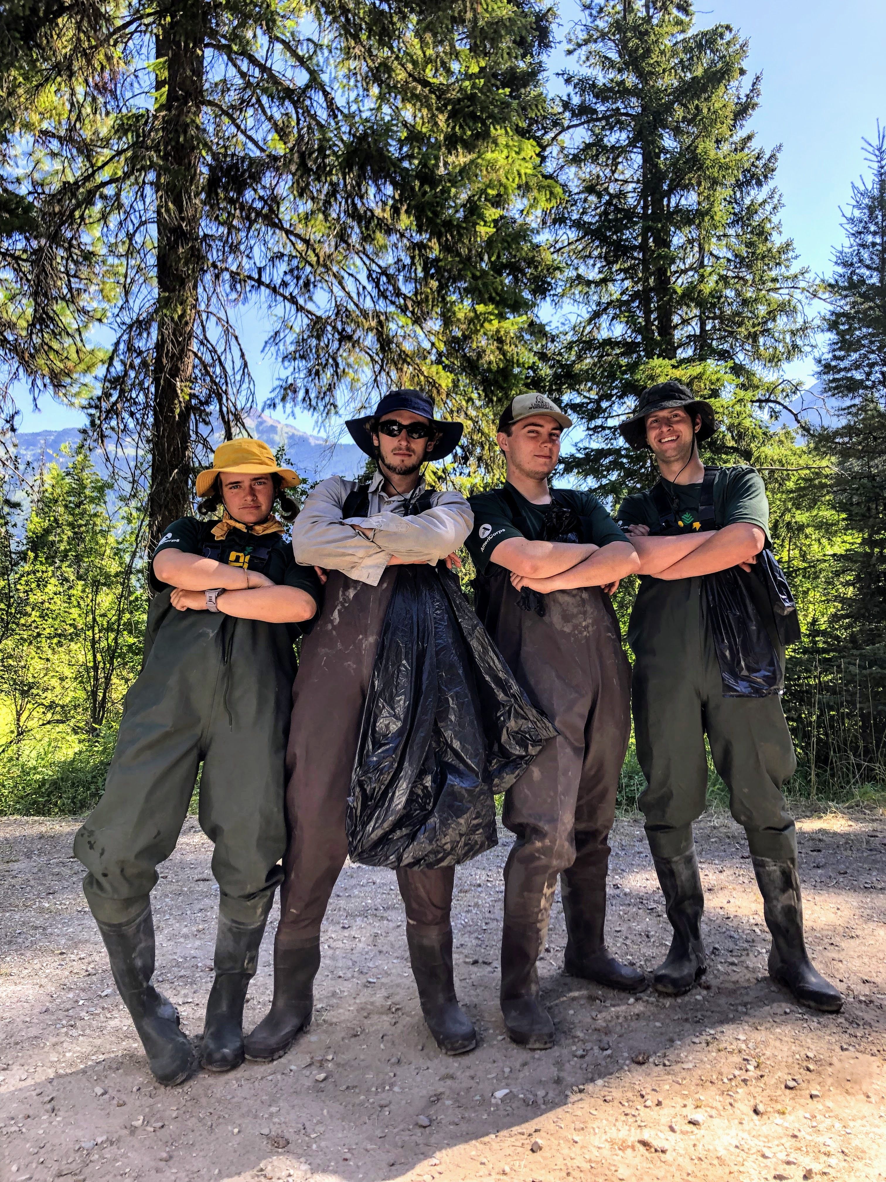 Four people wearing overall, high waders and holding trash bags pose in front of a lake.