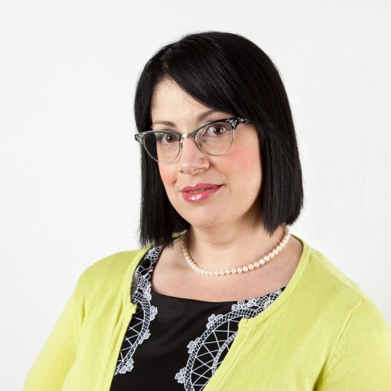 Sara Jane's headshot. She is facing to the right, and looking forward. She is wearing a black shirt, silver necklace, and yellow cardigan.