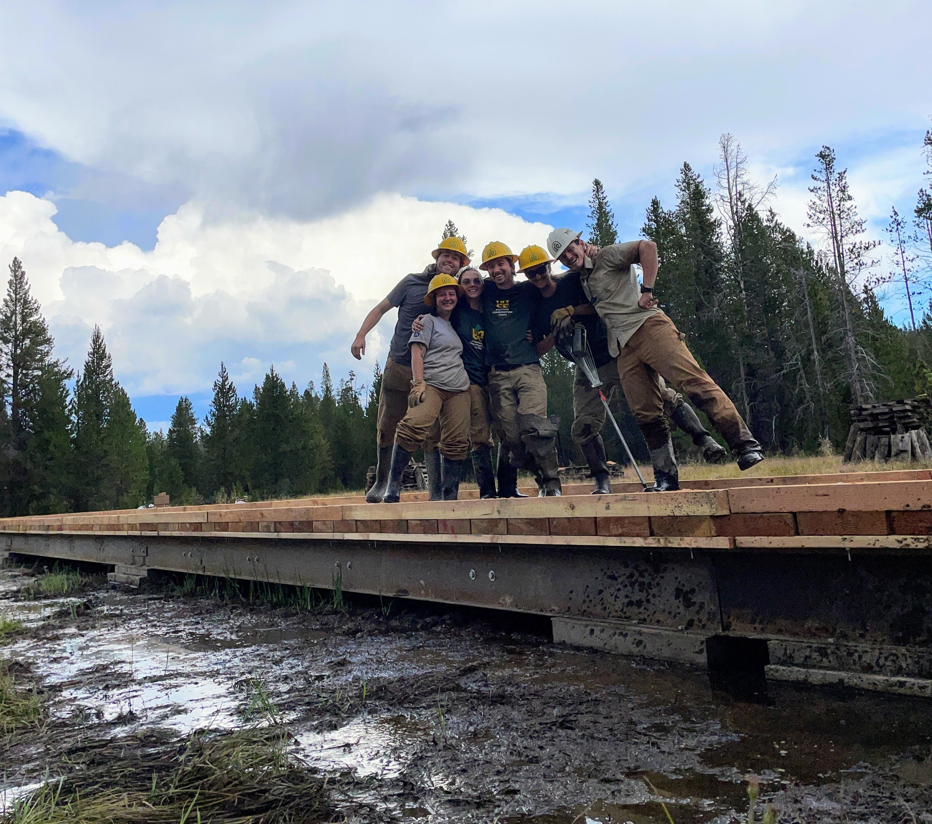 A crew poses on a long, steel and wood bridge they built that spans the length of a marsh.