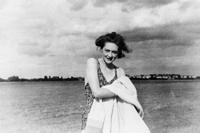A young woman smiles as she holds a towel and stands on a beach.