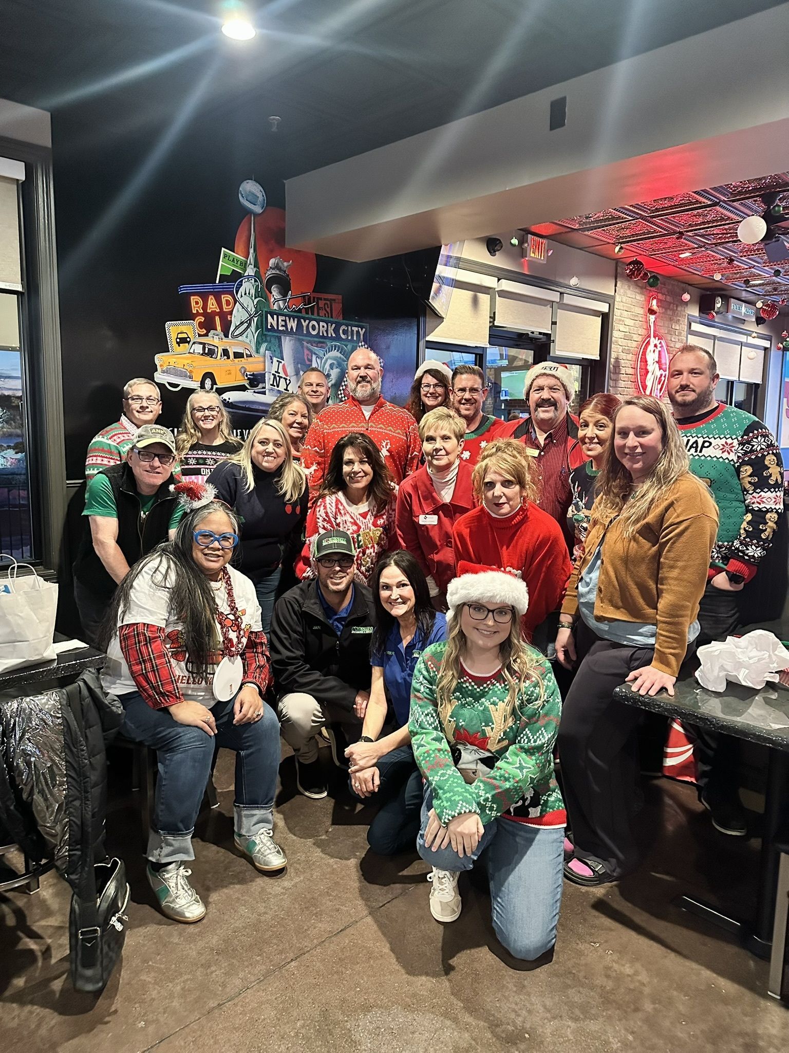 Group of people in festive holiday sweaters posing together indoors at a decorated event space.