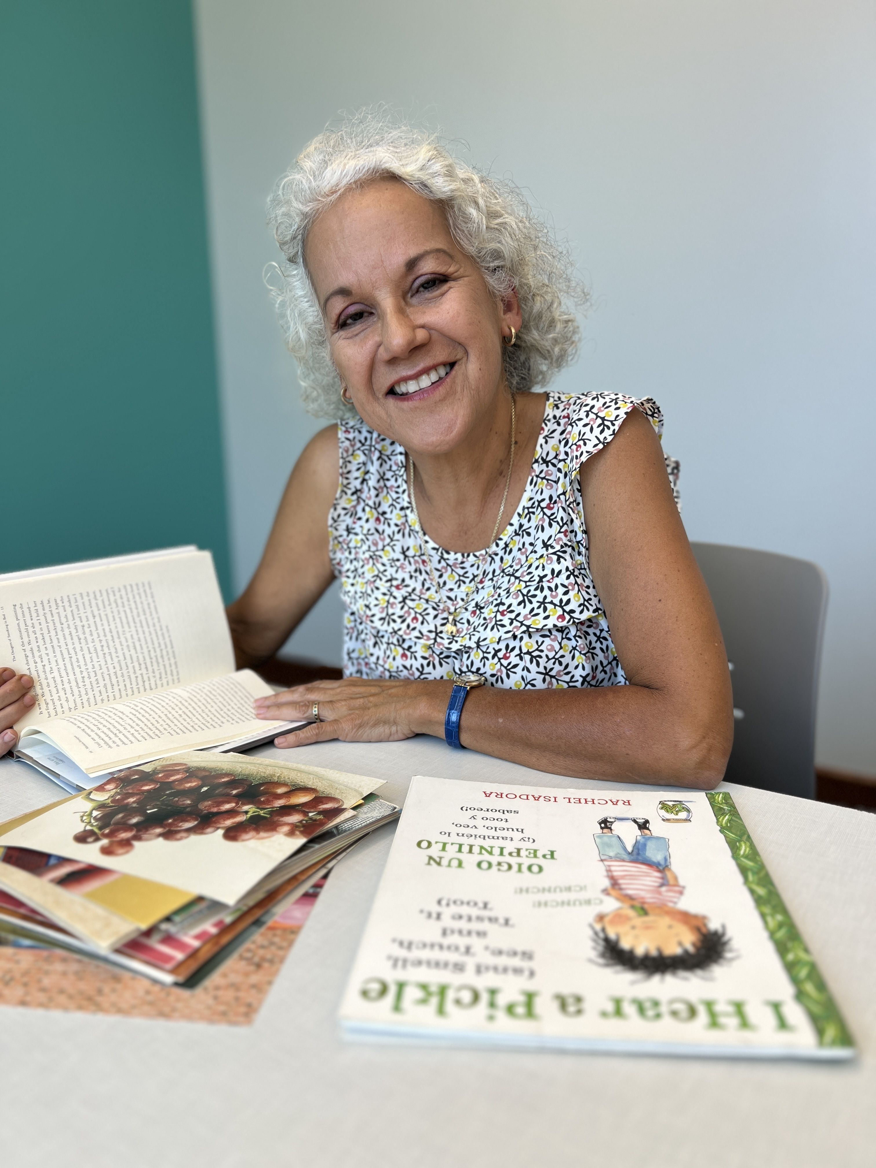 Margarita Patton smiles while sitting at a table in the library, surrounded by open books including a Spanish-language title and a children’s book titled I Hear a Pickle. She wears a floral sleeveless top and appears engaged and joyful, reflecting her lov