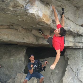 Alex Honnold holds on to a roof with one hand. A camera person films from below
