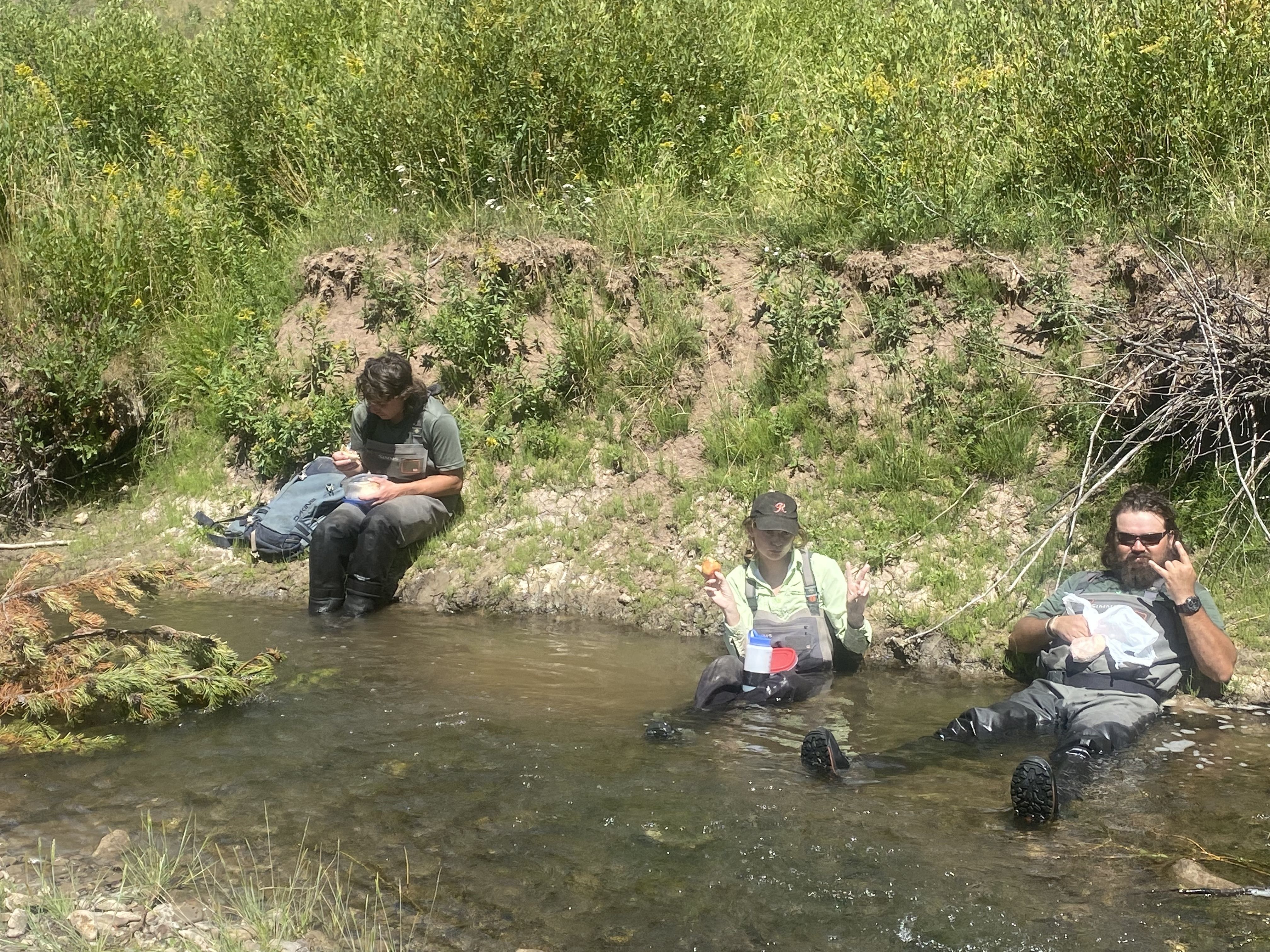 A crew sits in a creek eating their lunch.