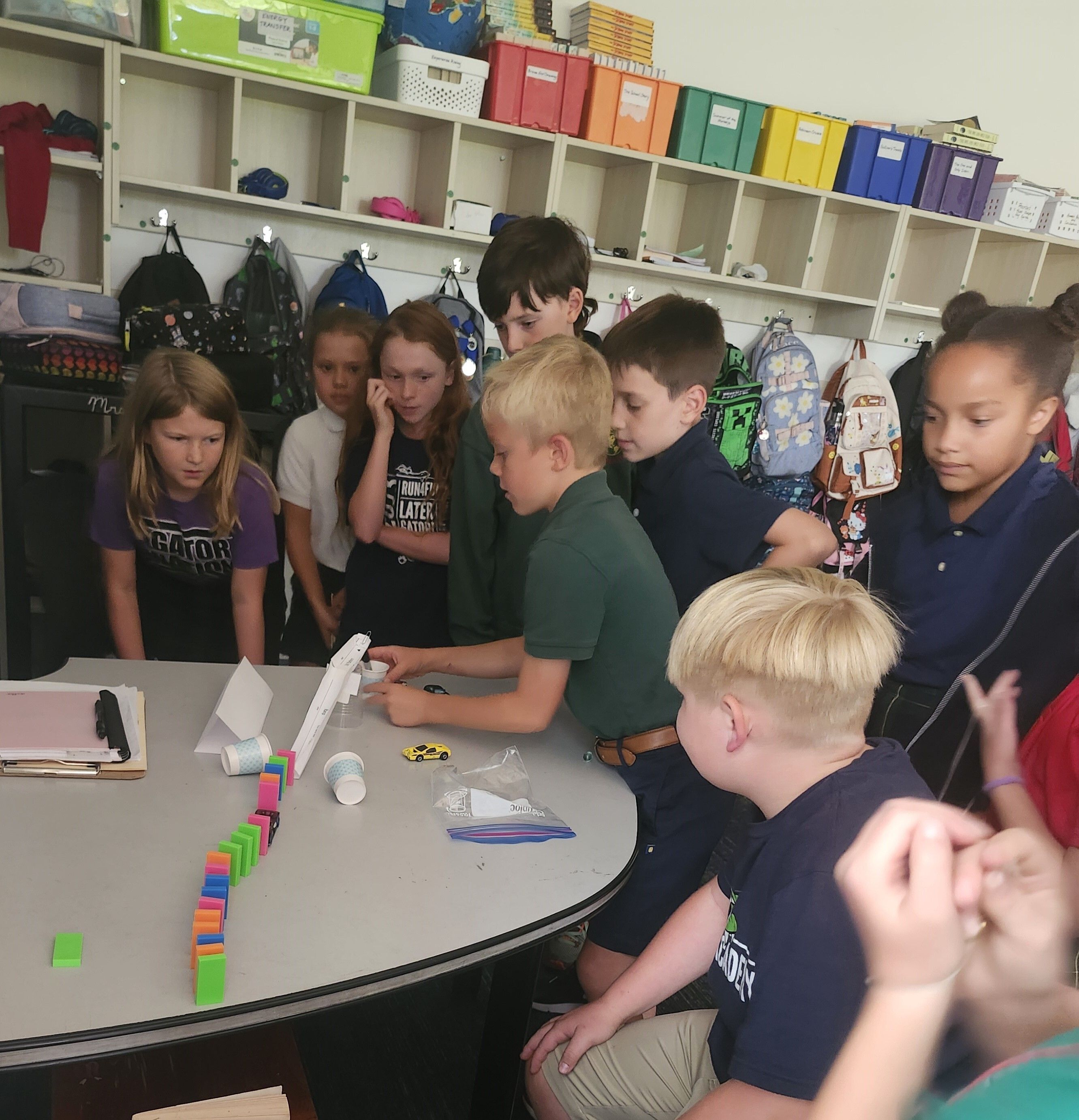 A group of elementary students gathered around a classroom table watch intently as a classmate demonstrates a hands-on science activity using dominoes, cups, and simple materials, while colorful bins and backpacks line the shelves behind them.
